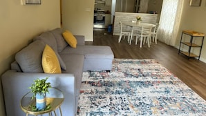 A living room with a grey sofa, a small glass table with a plant, and a colorful rug. There is a kitchen in the background with white cabinets and a dining area with white chairs. - Charming 2-bedroom house in Flowood, Pet Friendly w/Fenced Backyard and Sunroom (Brandon)