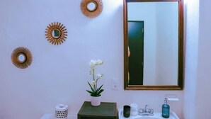 Une salle de bain avec un grand miroir, des petits miroirs décoratifs en forme de soleil fixés au mur, une plante en pot, une petite table avec des articles de toilette et un distributeur de serviettes.