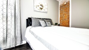 A bedroom with a large bed, grey and white bedding, a decorative wall piece, a brick accent wall, and sheer curtains. - Modern 1Br 1Ba Apartment Close Falls And Casino (Niagara Falls)