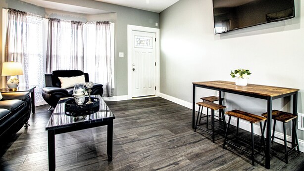 A living room with a black leather sofa, a coffee table, and a dining area with a wooden table and bar stools. - Modern 1Br 1Ba Apartment Close Falls And Casino (Niagara Falls)
