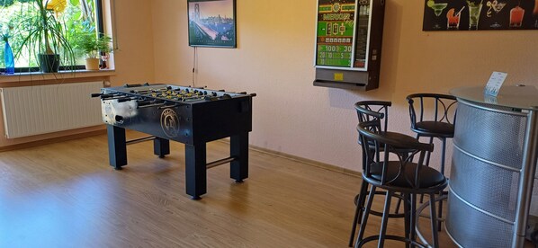 Ein Tischkicker in einem Raum mit Holzboden, einem Fenster mit Pflanzen, einem Wand-Fernseher und einer Bar mit Hockern.