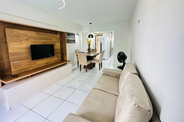 A modern living room with a flat-screen TV mounted on a wooden wall, a beige sofa, a dining area with a table and chairs, and a kitchen in the background. - Maison Escorel - LikeHome Hospedagens (Joao Pessoa)