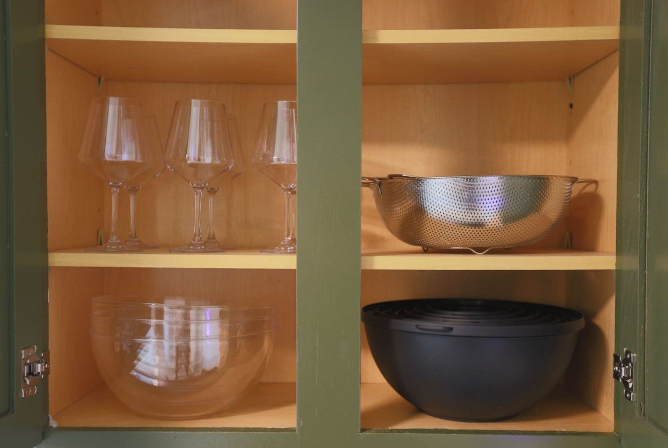 Un armario de madera con tres estantes. El estante superior sostiene tres copas de vino transparentes. El estante central contiene un colador de metal. El estante inferior tiene un bol grande transparente y un bol negro.
