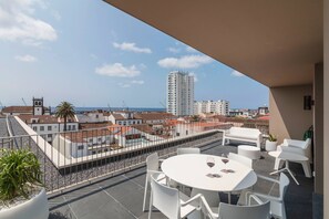 Eine Dachterrasse mit einem runden Tisch und weißen Stühlen, mit Blick auf eine Stadtlandschaft mit Gebäuden und blauem Himmel.