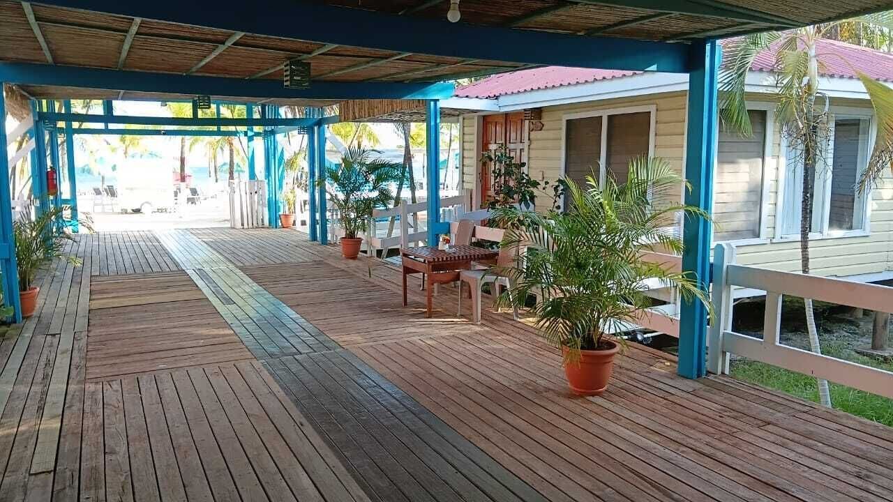 Une terrasse en bois couverte, agrémentée de plantes en pot, de meubles d’extérieur et offrant une vue sur une piscine et des bâtiments.