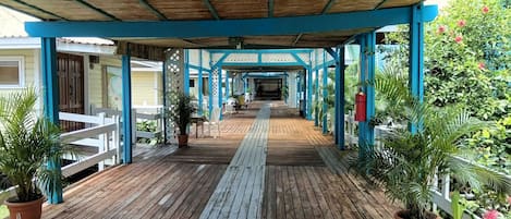A covered wooden deck with potted plants, white railings, and a view of a building in the background.