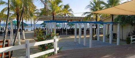 A wooden deck with a white fence, palm trees, and a covered area with tables and chairs.