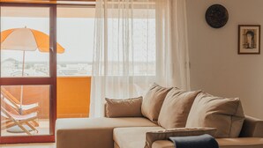 Ein Wohnzimmer mit einem beigefarbenen Sofa, einem großen Fenster mit durchsichtigen Vorhängen und Blick auf die Stadt.