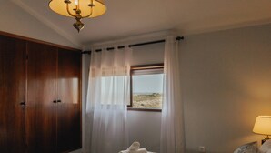 Ein Schlafzimmer mit großem Kleiderschrank, einem Bett mit zusammengelegtem Handtuch, einem Fenster mit Blick auf den Strand und einer Nachttischlampe.