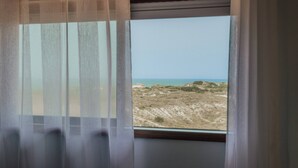 Ein Fenster mit durchscheinenden Vorhängen, das Blick auf eine Strandlandschaft mit Dünen und dem Meer bietet.