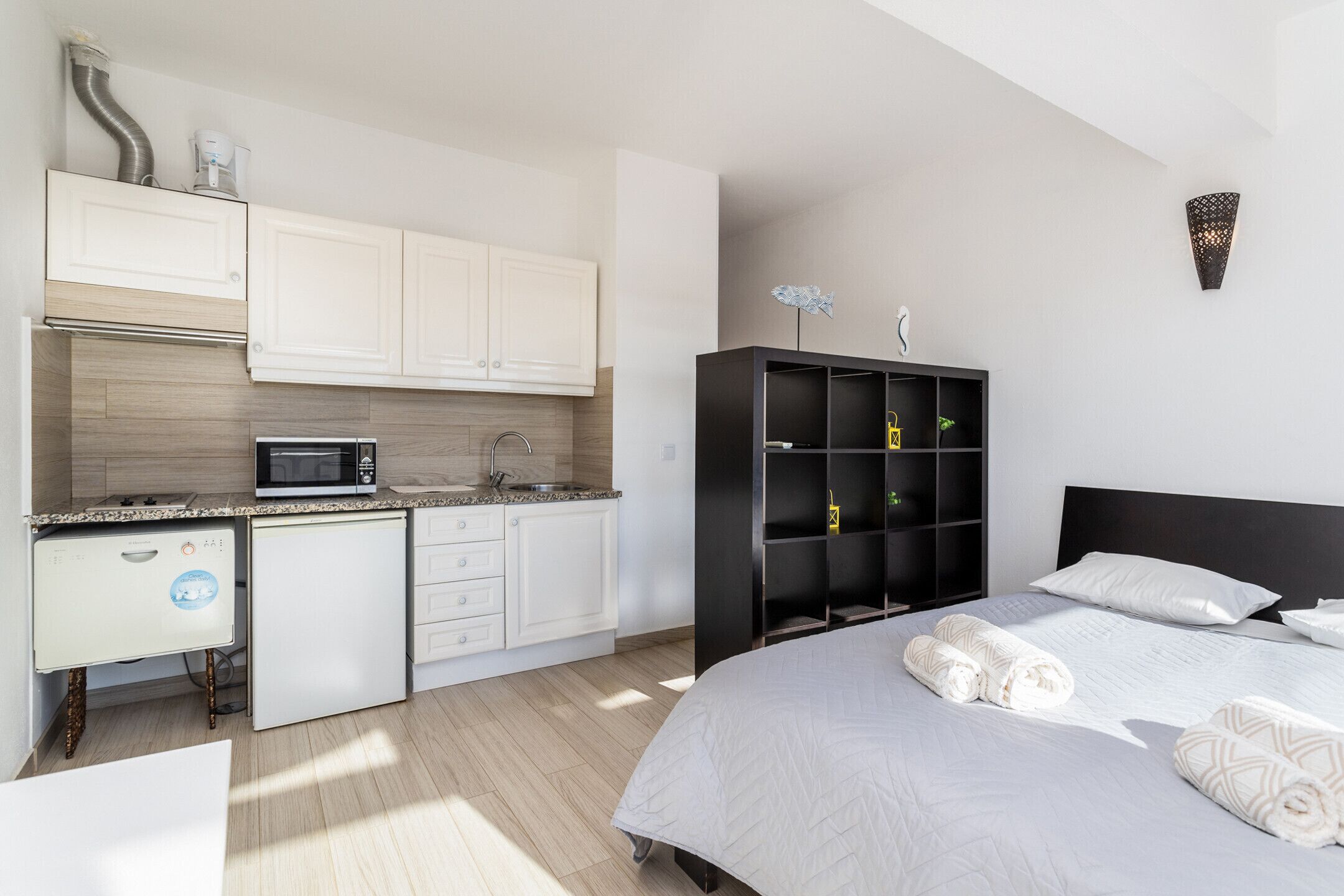 Quarto de hotel moderno com cozinha compacta, cama com toalhas impecáveis e um armário preto na parede.