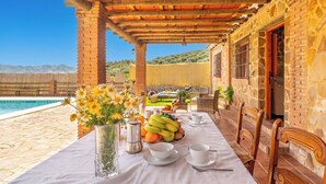 Ein Esstisch ist reichhaltig mit Früchten, Tassen und einem Blumenstrauß gedeckt, mit Blick auf einen Pool und eine gebirgige Landschaft.