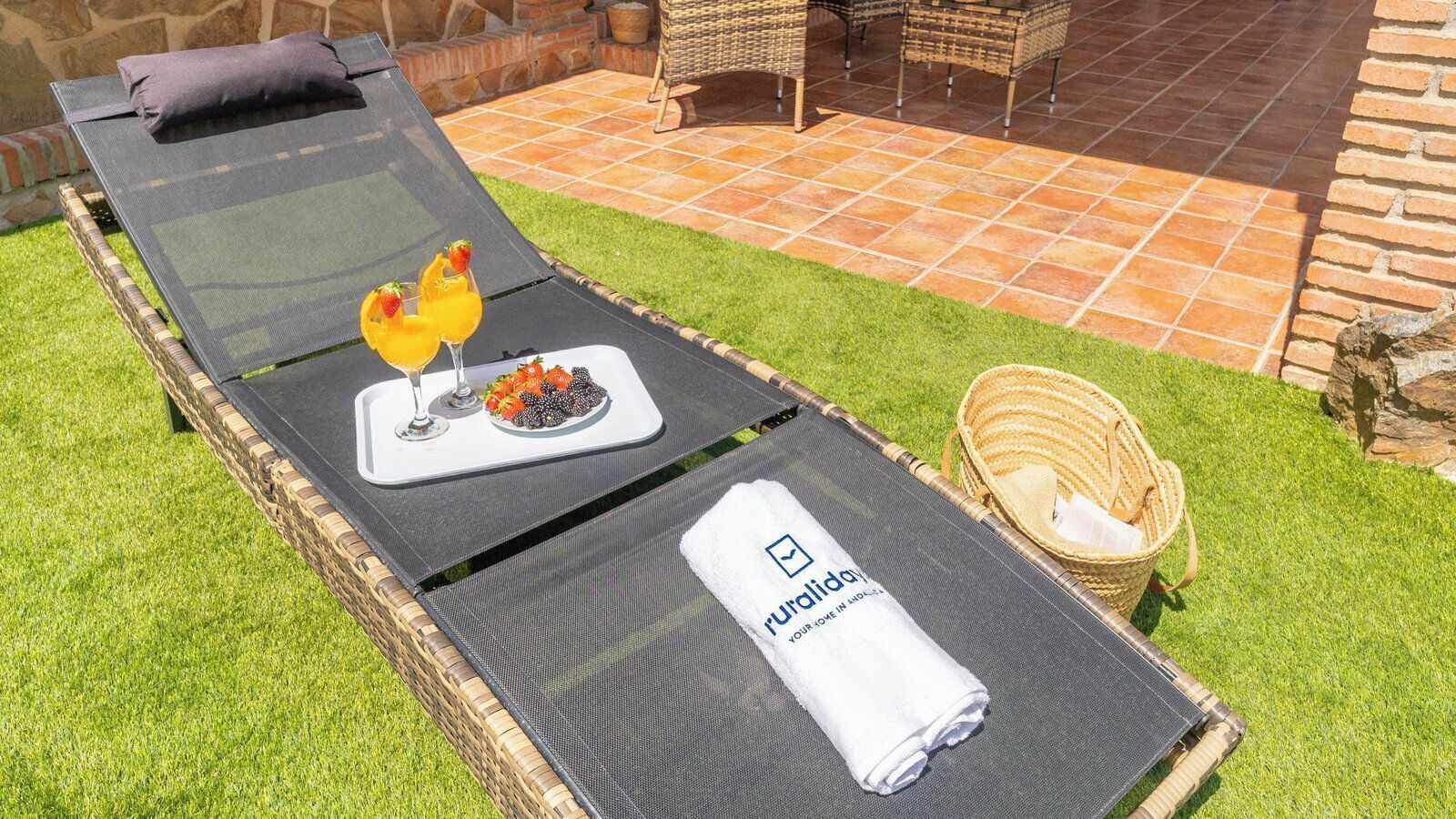 Une chaise longue avec un plateau de fruits et une boisson, une serviette ornée d’un logo, et des meubles en osier en arrière-plan.
