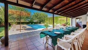 An outdoor dining area with a pool, covered by a wooden roof, and surrounded by greenery.