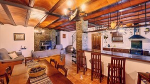 A cozy living room with a fireplace, wooden furniture, and a kitchen area with a stove and hanging lights.