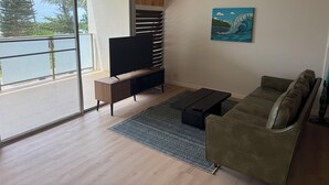 A modern living room with a wooden ceiling, a large sliding glass door to a balcony, a flat-screen TV, a green sofa, and a patterned rug. - Tranquil 1-bedroom condo on a sandy beach in Hauula (Hauula)