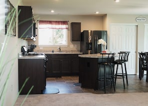 A kitchen with dark cabinetry, a stainless steel refrigerator, a coffee machine, and a window with a red valance. There are three black bar stools and a white door labeled "PANTRY".
