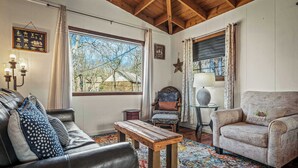 A cozy living room with a leather sofa, a wooden coffee table, and a patterned rug. There is a window with curtains, a star decoration on the wall, and a lamp on a side table. - Mountain Treehouse - Magpie Nest (Mentone)