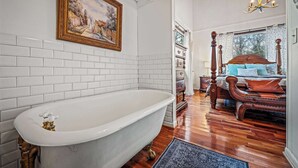A room with a white bathtub, wooden floor, a bed with a headboard, and a painting on the wall. - Mountain Treehouse - Magpie Nest (Mentone)