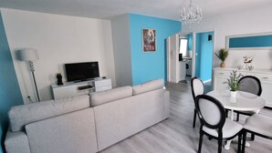 A modern living room with a grey sofa, a flat-screen TV, a dining table with chairs, and a chandelier.