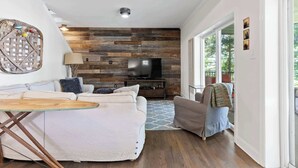 A modern living room with a wooden wall panel, a flat-screen TV, a sofa, a dining table, and a view through large windows.