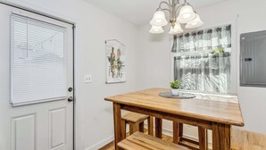 Un coin repas avec une table et des bancs en bois, un lustre, une fenêtre avec des rideaux légers et une porte avec des stores.