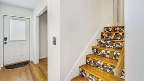 Un escalier avec des marches en bois et des contremarches recouvertes de mosaïques, une porte blanche avec un paillasson, et un mur de couleur claire.