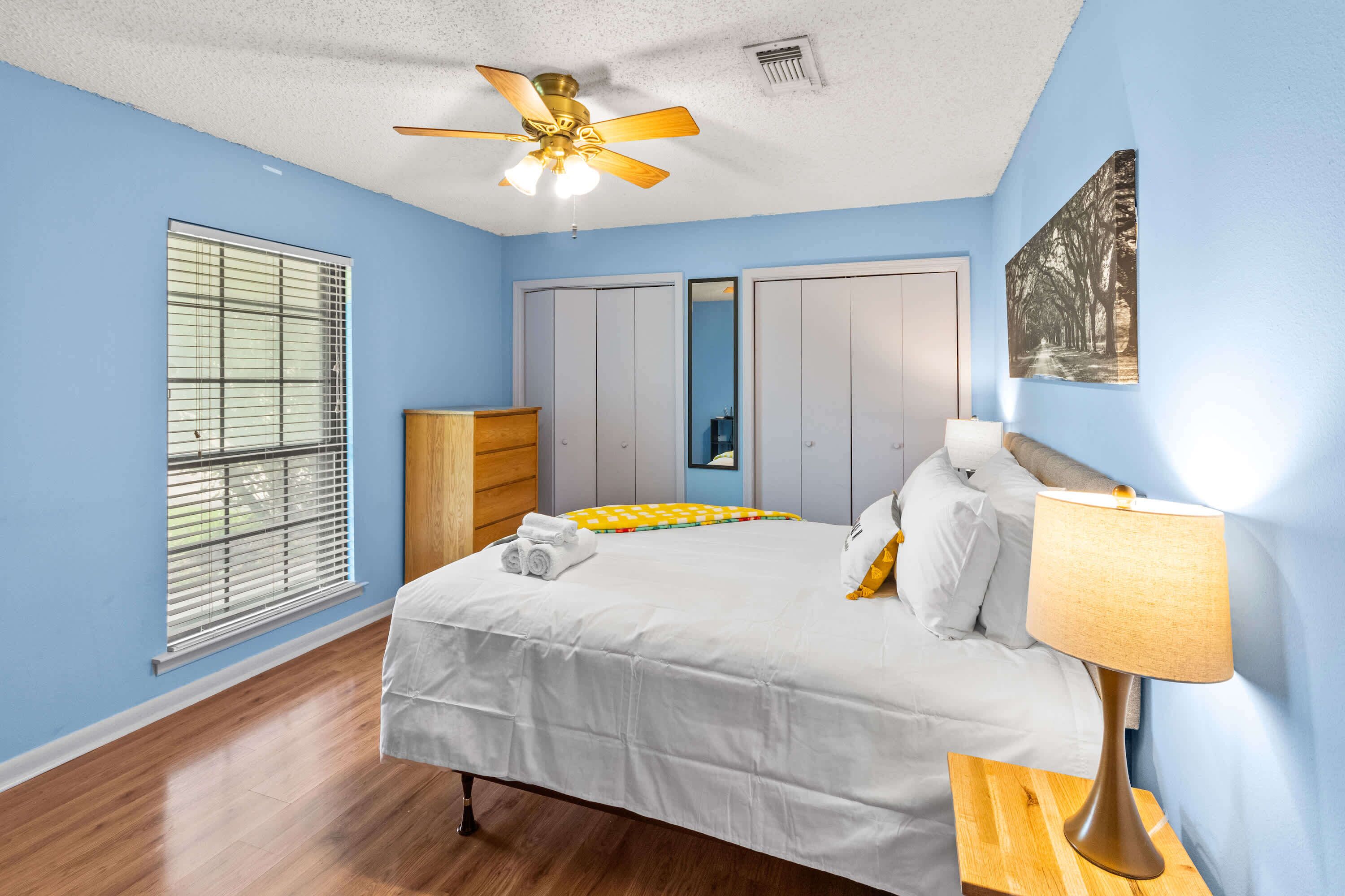 A bedroom with a bed, a ceiling fan, a bedside table with a lamp, a wardrobe, and a window with blinds.
