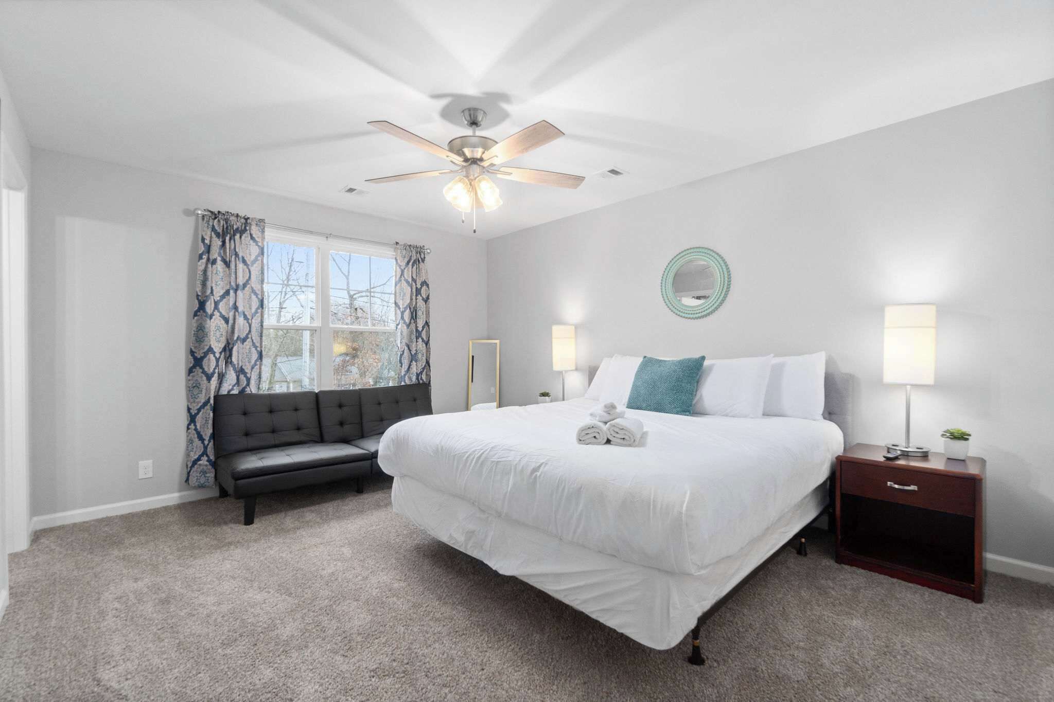 Une chambre à coucher avec un grand lit, un ventilateur de plafond, une table de chevet avec une lampe, un banc et une fenêtre agrémentée de rideaux à motifs.