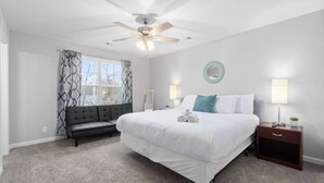 A bedroom with a large bed, a ceiling fan, a nightstand with a lamp, a bench, and a window with patterned curtains.