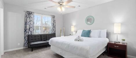 A bedroom with a large bed, a ceiling fan, a nightstand with a lamp, a bench, and a window with patterned curtains.