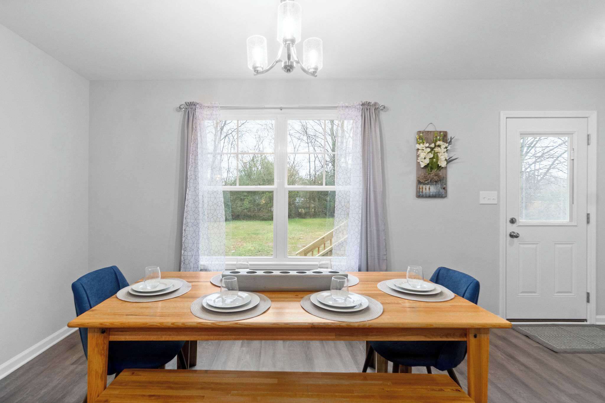 Un coin repas avec une table en bois dressée pour quatre personnes, un lustre et une vue sur un jardin, visible à travers une grande fenêtre.