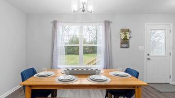 Un coin repas avec une table en bois dressée pour quatre personnes, un lustre et une vue sur un jardin, visible à travers une grande fenêtre.