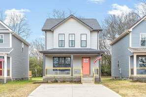 A two-story house with a red door, a small porch with a yellow railing, and an American flag on a flagpole.