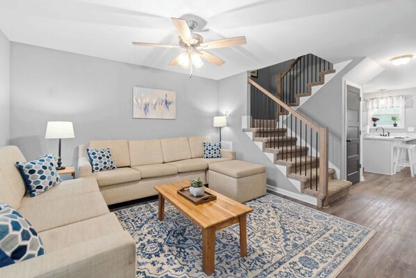 A living room with a beige sofa, a wooden coffee table, and a patterned rug. A staircase with wooden railings leads to a kitchen area.