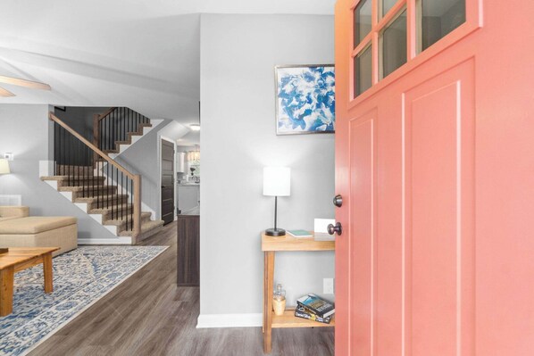 A modern interior with a staircase, a wooden table, a blue rug, a red door, and a wall-mounted lamp.