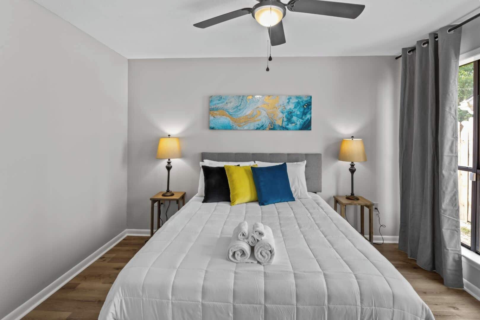 Un dormitorio con una cama, dos mesitas de noche, un ventilador de techo, una ventana con cortinas y un cuadro en la pared.