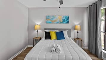 Un dormitorio con una cama, dos mesitas de noche, un ventilador de techo, una ventana con cortinas y un cuadro en la pared.