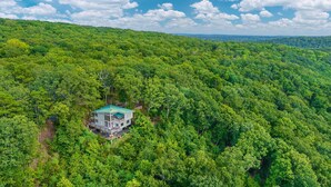 A large, two-story house surrounded by dense forest with a clear view of the sky.