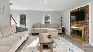A living room with a sectional sofa, a coffee table, a fireplace, and a TV mounted on a brick wall. - Near NC State Campus & Downtown - The Maple Marvel (Raleigh)