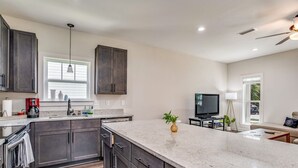 A modern kitchen with dark cabinets, a large island, and a TV in the living area. - Near Downtown, Dining & Base - Beach Flow Bungalow (Pensacola)