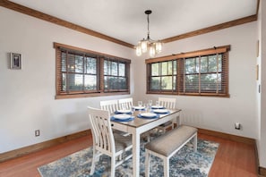 Uma área de refeições com mesa de madeira posta para quatro pessoas, cadeiras brancas e um banco. Há duas janelas com persianas de madeira, um candeeiro e um quadro na parede.