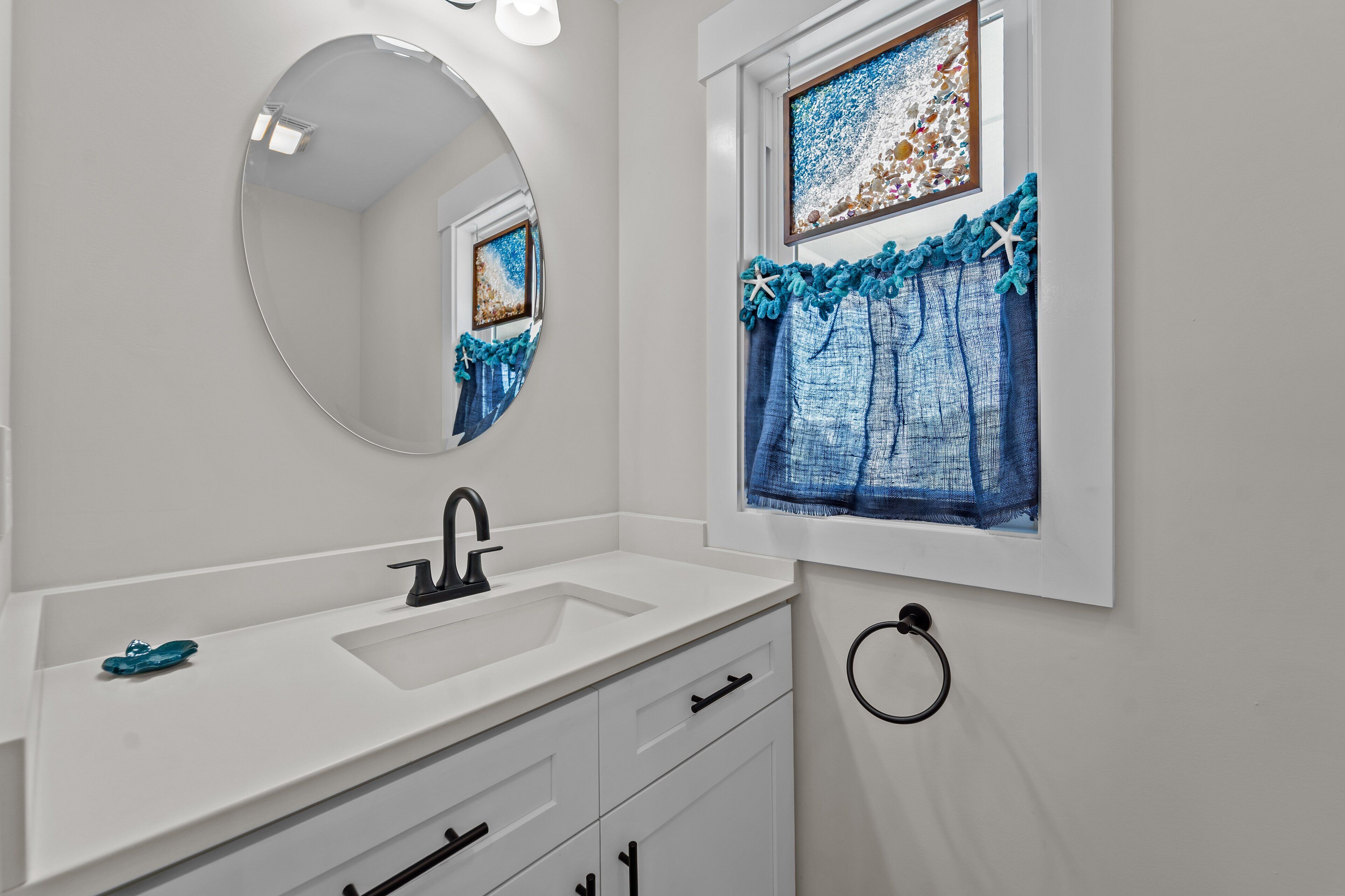 Un baño con un lavabo blanco, un espejo redondo y una ventana con cortina azul.
