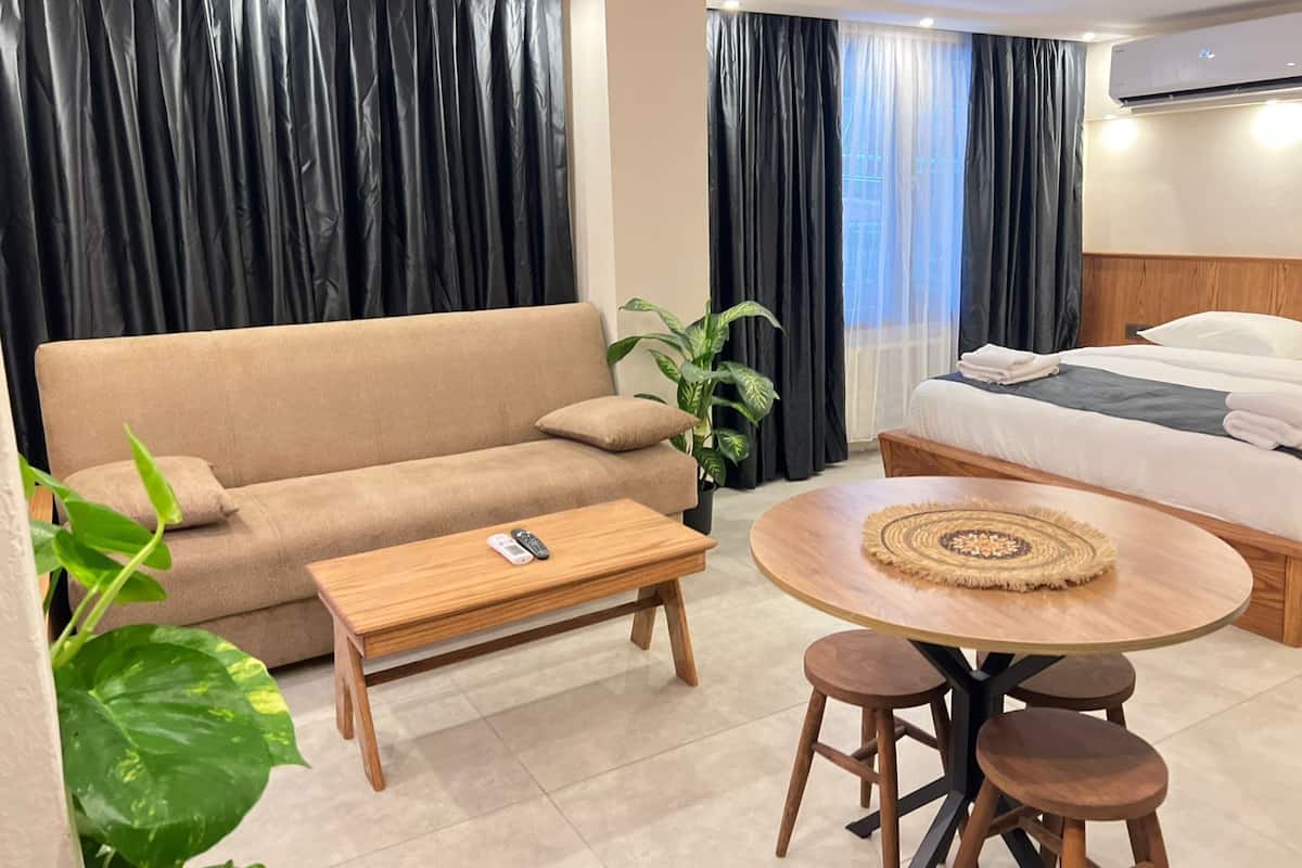 A modern hotel room with a sofa, a round wooden table with stools, a bed with a white duvet, and a potted plant.