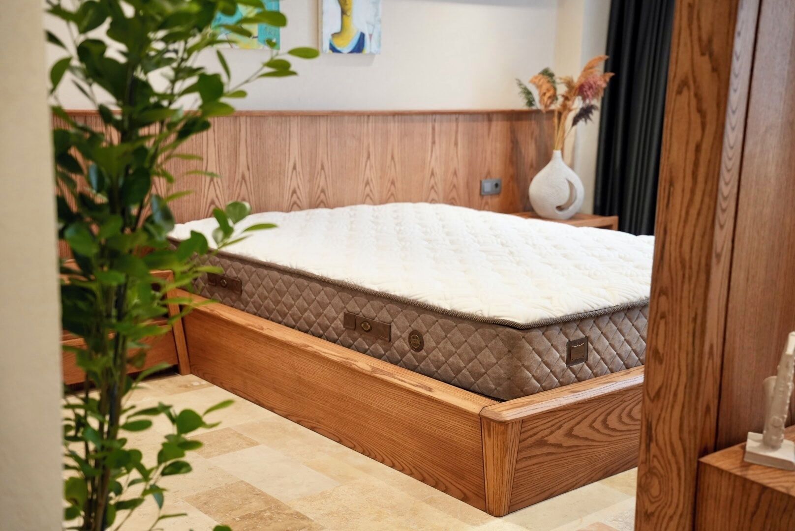 Un lit en bois avec un matelas matelassé, dans une pièce dotée d’une tête de lit en bois et d’un vase contenant des fleurs séchées, posé sur une table de chevet.