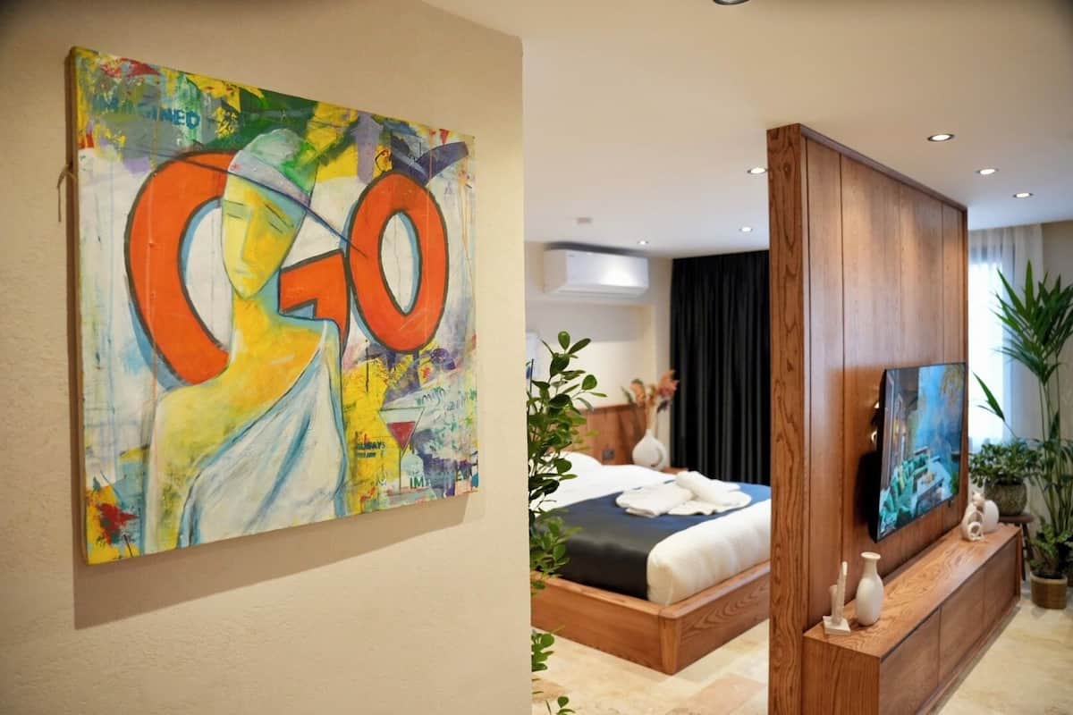 A modern hotel room with a large abstract painting, a bed with a blue and white bedspread, and a wooden console table with a television and decorative items.