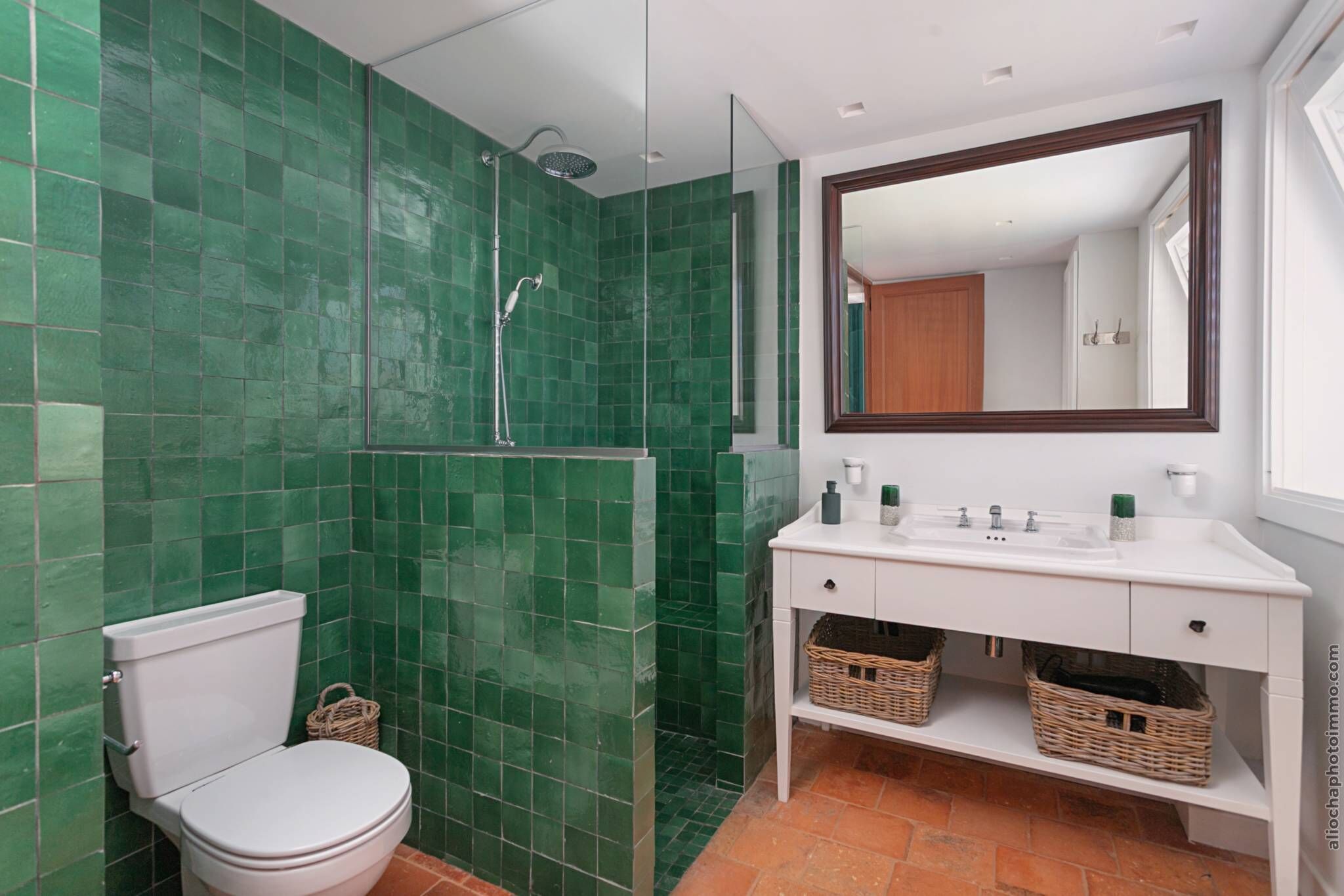 A bathroom with green tiles, a white toilet, a white sink with a mirror above it, and wicker baskets under the sink.