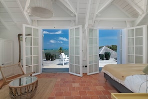 A bedroom with a large bed, a wicker chair, a glass table, and a view of the ocean through open French doors. - Les Frangipaniers, an extraordinary villa with a pool and direct access to the ocean   (Unknown)