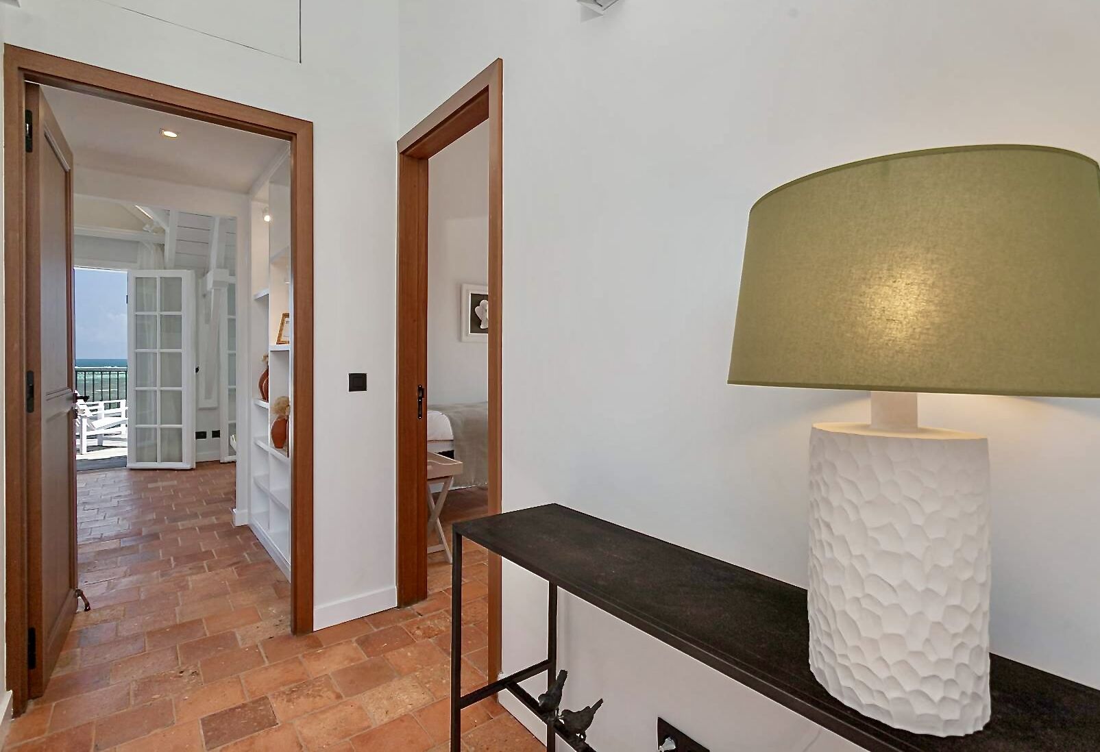Un couloir avec une porte en bois donnant sur un balcon, un mur blanc orné d’une lampe décorative, et un sol carrelé.