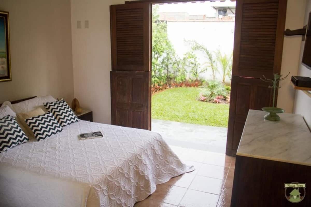 Un dormitorio con cama, mesita de noche, cómoda y vista a un jardín a través de puertas abiertas.